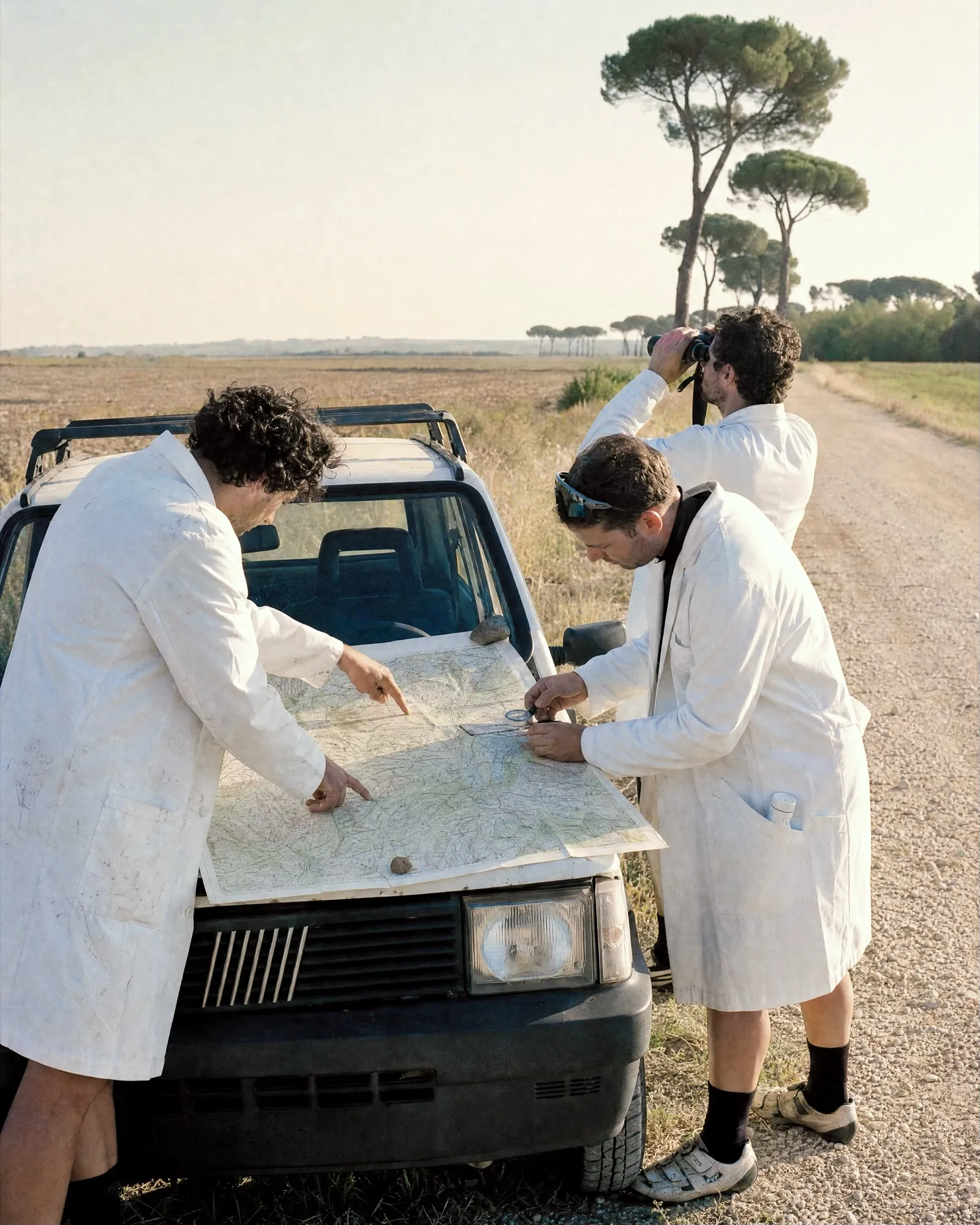 Tre soci in camice bianco leggono una mappa sul cofano di una Panda su una strada bianca fra i pini di Maccarese